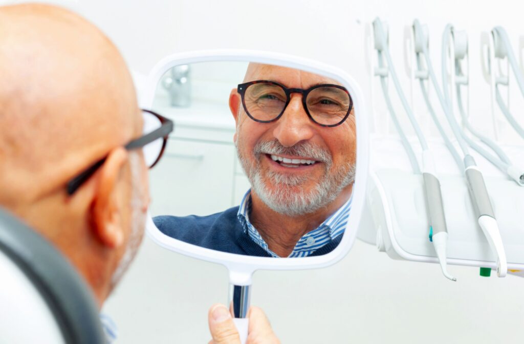  An older man wearing glasses smiles while looking at his teeth in a handheld mirror during a dental visit, with dental instruments visible in the background