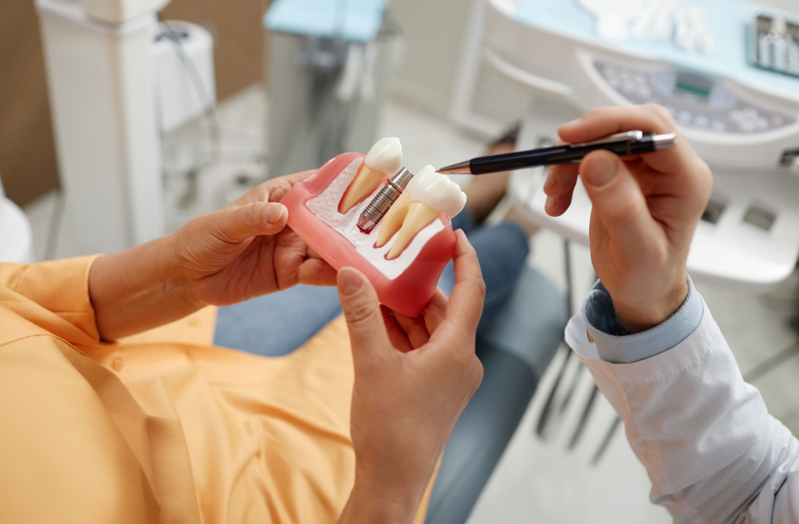 A dental professional explains a dental implant using a jaw model, pointing to a metal implant post placed between two teeth during a patient consultation